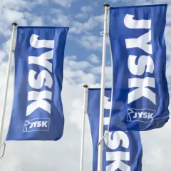 Niebieskie flagi z białym logo JYSK na tle błękitnego nieba z chmurami.