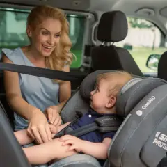 Lächelnde Mutter sichert ihr Baby im Kindersitz vorne im Auto.