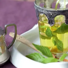 Ein Glas mit frischem Minztee, daneben eine Teekanne aus Metall.