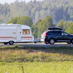 Ciemny SUV ciągnie białą przyczepę kempingową po drodze, na tle lasu.