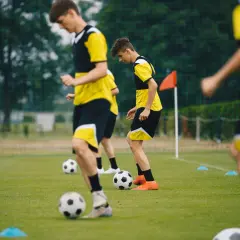 Młodzi piłkarze w żółto-czarnych strojach ćwiczą na boisku. Widać piłki i pachołki, co sugeruje trening dla różnych kategorie wiekowe w piłce nożnej.