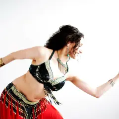 Kobieta w stroju do tańca brzucha, z błyszczącym topem i czerwoną spódnicą, pokazuje, jak zacząć przygodę z tym tańcem.