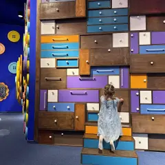 Dziewczynka odkrywa tajemnice w kolorowej ścianie szuflad, jak w Akademii Pana Kleksa w Katowicach.