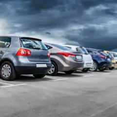 Rząd samochodów na parkingu, w tym jeden z włoską rejestracją, pod burzowym niebem.