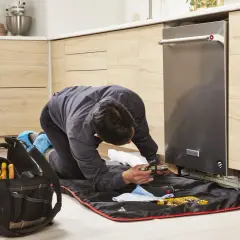 A technician kneels to replace a dishwasher, surrounded by tools and a protective mat.