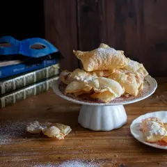 Chiacchiere friabili e bollose, cosparse di zucchero a velo, su un piatto con una maschera veneziana e libri antichi sullo sfondo.