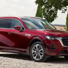Lśniąca, czerwona Mazda CX-80 zaparkowana na żwirowej drodze, otoczona zielenią i ceglanym murem.