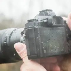 Dłoń trzyma aparat z obiektywem, pokryty kroplami deszczu. To była trudna, ale udana sesja w deszczu.