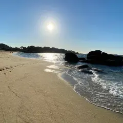 La **playa de Tulla** bajo un sol radiante. Huellas en la arena dorada y el mar azul acarician las rocas oscuras.