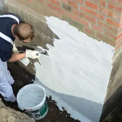 Mann dichtet mit Spachtel und Mörtel die Gartenmauer ab. Ein Eimer mit Abdichtungsmaterial steht bereit.