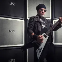Mężczyzna w czapce i okularach gra na gitarze elektrycznej przed ścianą wzmacniaczy. Wygląda jakby był gotowy na koncert dla fanów popcorn acid drinkers.