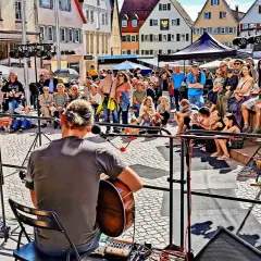 Ein Musiker spielt Gitarre, während eine Sängerin auf einer Bühne steht. Viele Menschen sitzen und stehen davor, um die **Veranstaltung weil der Stadt** zu genießen.
