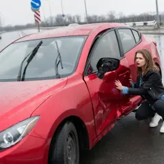 Młoda kobieta ogląda uszkodzone drzwi czerwonego samochodu po wypadku. Widać wgniecenie i urwane lusterko. Zawieszenie auta mogło ucierpieć.