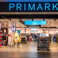 Wejście do sklepu Primark w Polsce. Widać neonowy napis "PRIMARK" i wystawione ubrania na manekinach i wieszakach.