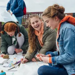 Drei Mädchen sitzen im Freien und malen auf einer großen Papierrolle. Sie lachen und arbeiten zusammen, als ob es eine kleine **Abschlussfeier** wäre.