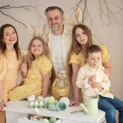 Rodzinna sesja zdjęciowa z zajączkami na głowach. Uśmiechnięci rodzice i dzieci w żółtych strojach pozują na tle wiosennych dekoracji.