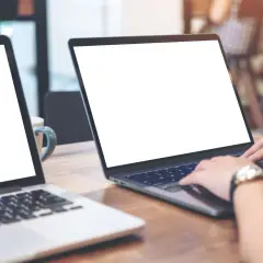 Zwei Laptops auf einem Holztisch. Eine Person nutzt einen Laptop als Monitor, während die Hände auf der Tastatur liegen.