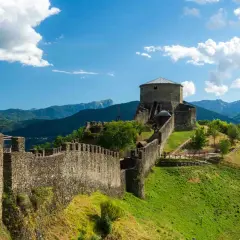 Roccia imponente e mura antiche del **San Romano in Garfagnana** si stagliano su un paesaggio verdeggiante, sotto un cielo azzurro con nuvole bianche.
