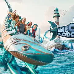 Un train de montagnes russes en forme de requin mécanique sur l'eau, avec un phare et un village de pêcheurs.