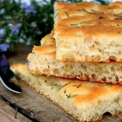 Gestapelte Focaccia-Stücke mit Rosmarin, die an das Backen von Pizza erinnern. Ein Messer liegt daneben auf einem Holzbrett.