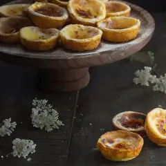 Pasteles de Belém recién horneados, dorados y cremosos, espolvoreados con azúcar glas sobre un soporte de madera.
