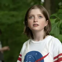 Dziewczynka w koszulce NASA patrzy w górę, jakby chciała zostawić świat za sobą. W tle niewyraźna postać.