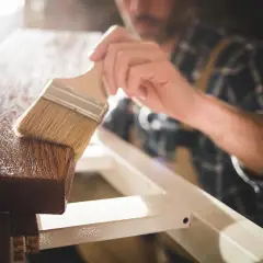 Un falegname applica una finitura protettiva su un pezzo di legno. Dopo l'impregnante, cosa si mette? Una vernice trasparente o una cera per esaltare la venatura.