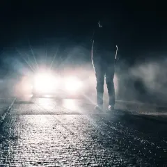 Silhouette d'une personne sur une route mouillée, éclairée par un appel de phare éblouissant dans la nuit brumeuse.