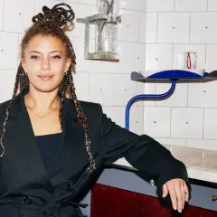 Matilda Jackson, med flätor uppsatta i en knut, lutar sig mot en bardisk med flaskor i bakgrunden.