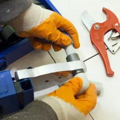 Ręce w rękawiczkach montują elementy systemów łączenia rur wodnych przy użyciu zgrzewarki. Obok leżą czerwone nożyce do rur.