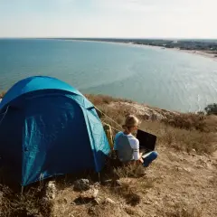 Niebieski namiot i osoba z laptopem na wzgórzu, z widokiem na spokojne, lazurowe morze. Idealny camping lazurowe wybrzeże.