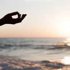 Dłoń w geście mudry na tle zachodzącego słońca nad morzem. Chwila głębokiej mindfulness i spokoju.