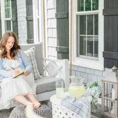 A woman embodies the coastal grandma aesthetic, writing in a notebook on a porch swing, with lemonade and hydrangeas nearby.