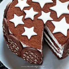 Torta Pan di Stelle ricoperta di cacao, decorata con stelle bianche e biscotti. Una fetta mostra gli strati di crema.