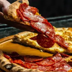 Kawałek pizzy pepperoni unoszony nad całą pizzą na drewnianej desce.