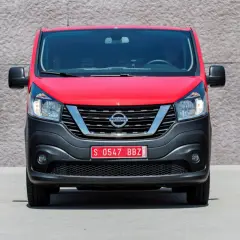 Czerwony Nissan NV300 z czarnym zderzakiem i chromowaną atrapą chłodnicy stoi przed betonową ścianą.