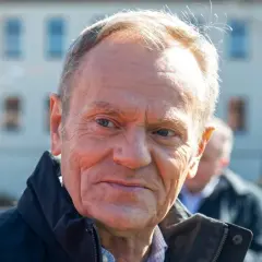 Portret Donalda Tuska w czarnej kurtce, z innymi osobami w tle.