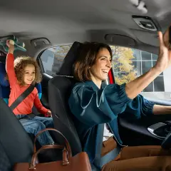 Glad mamma justerar backspegeln i sin Toyota, med barnet i bakgrunden som leker med en leksak. Med Toyota garanti kan du känna dig trygg.