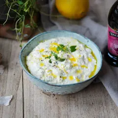 En skål med krämig, vispad fetaost toppad med olivolja och örter. Bredvid syns vitlök, citron och en flaska fond.