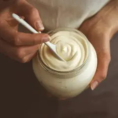 Mani che mescolano con un cucchiaino una vellutata maionese fatta in casa in un barattolo di vetro.