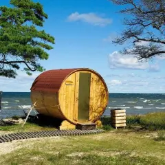 Beczka-sauna nad morzem, jedna z nietypowych atrakcji Juraty. Drewniana konstrukcja z czerwonym dachem, przy plaży, pod drzewami.