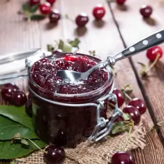Słoik pełen domowego musu wiśniowego, z łyżeczką gotową do degustacji. Wokół świeże wiśnie i liście.