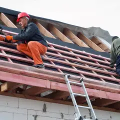 Dwaj budowlańcy montują wiatroizolację na dachu. Jeden z nich w pomarańczowych spodniach i czerwonym kasku wbija gwoździe.