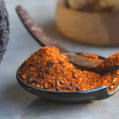 A dark, textured rock sits beside a small bowl filled with a vibrant 7 spice recipe blend. A wooden spoon rests in the bowl, ready to scoop the aromatic mixture.