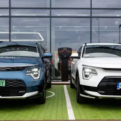 Dwa nowe samochody Kia Niro, jeden niebieski, drugi biały, stoją obok siebie na zielonym parkingu przed nowoczesnym budynkiem z dużymi oknami.
