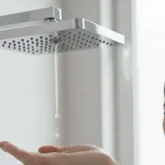 Une femme au visage déconfit teste l'eau de la douche. Elle n'a que de l'eau chaude dans sa douche, quelle déception !