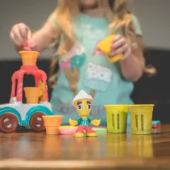 Dziecko bawi się w play doh warsztat, tworząc lodowe przysmaki. Na stole kolorowe foremki i figurka lodziarza.