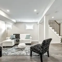 A bright, modern living area in a finished basement, featuring a white sectional, patterned rug, and a staircase leading up.