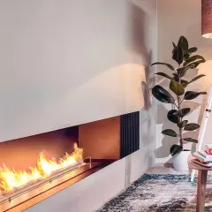 Nowoczesny kominek bio wolnostojący z płonącym ogniem, obok którego stoi lampa i roślina.