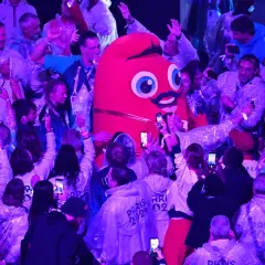 La Phryge JO, mascotte des JO, interagit avec une foule joyeuse. Les gens filment et prennent des photos avec leurs téléphones.